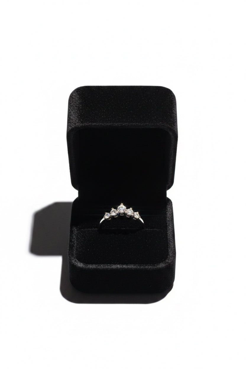 A silver ring with a small cluster of clear gemstones sits in an open black velvet ring box on a white background.