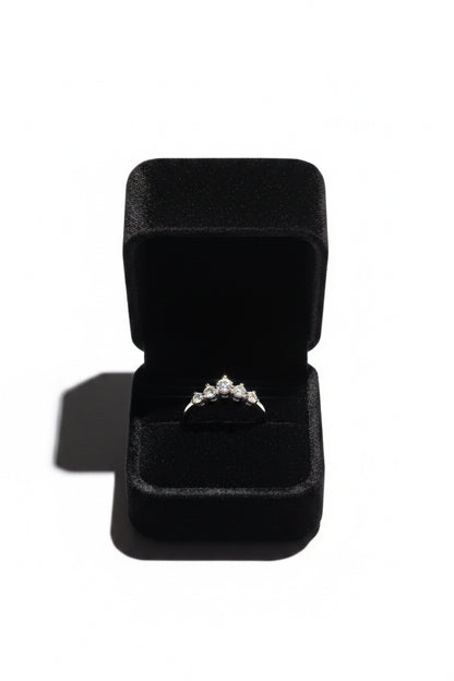 A silver ring with a small cluster of clear gemstones sits in an open black velvet ring box on a white background.