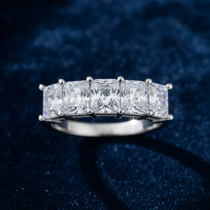A silver-tone ring featuring five rectangular clear gemstones in a row, set against a dark blue textured background.