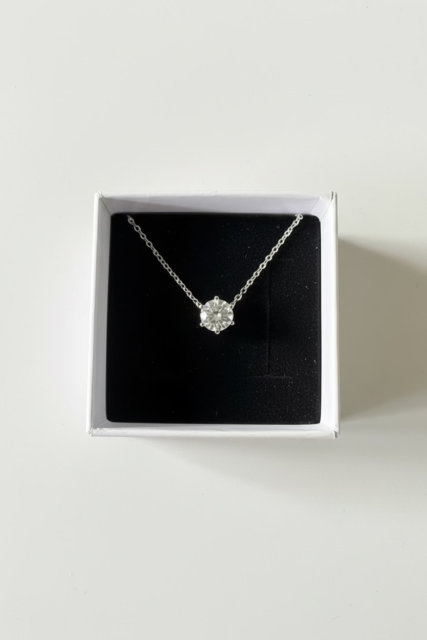 A silver-toned diamond pendant necklace displayed inside an open white jewelry box with a black velvet lining, set on a gray textured surface.