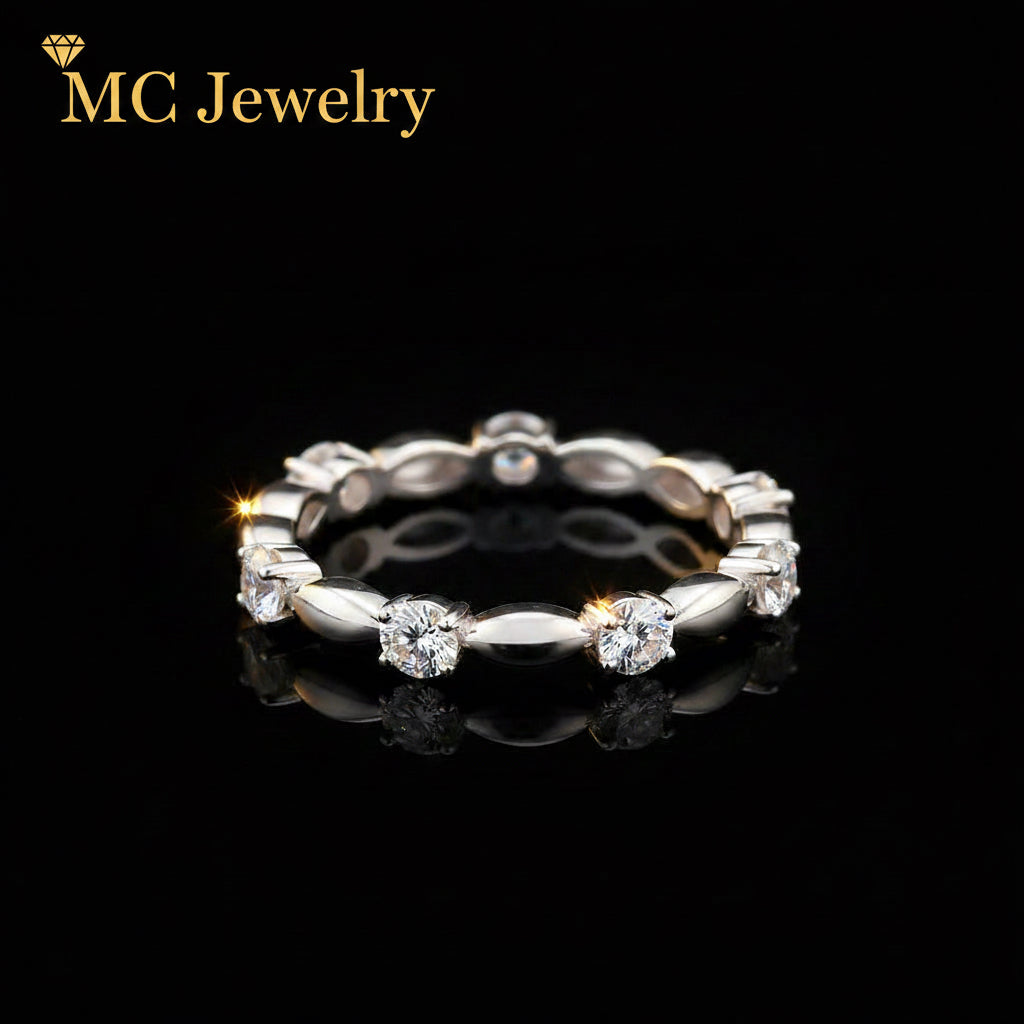A silver-toned eternity ring featuring round brilliant-cut diamonds set in a continuous band, displayed on a reflective black surface against a black background.