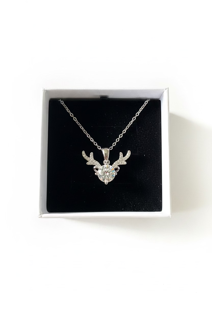 A silver-toned necklace with a reindeer head pendant set with clear stones, displayed inside a white jewelry box with a black velvet surface.