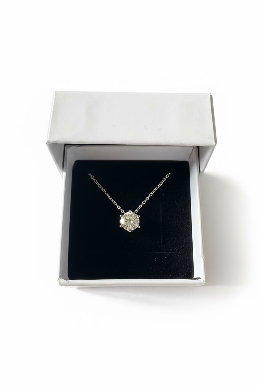 A silver-toned pendant necklace with a clear faceted gemstone displayed inside an open white jewelry box with a black velvet insert
