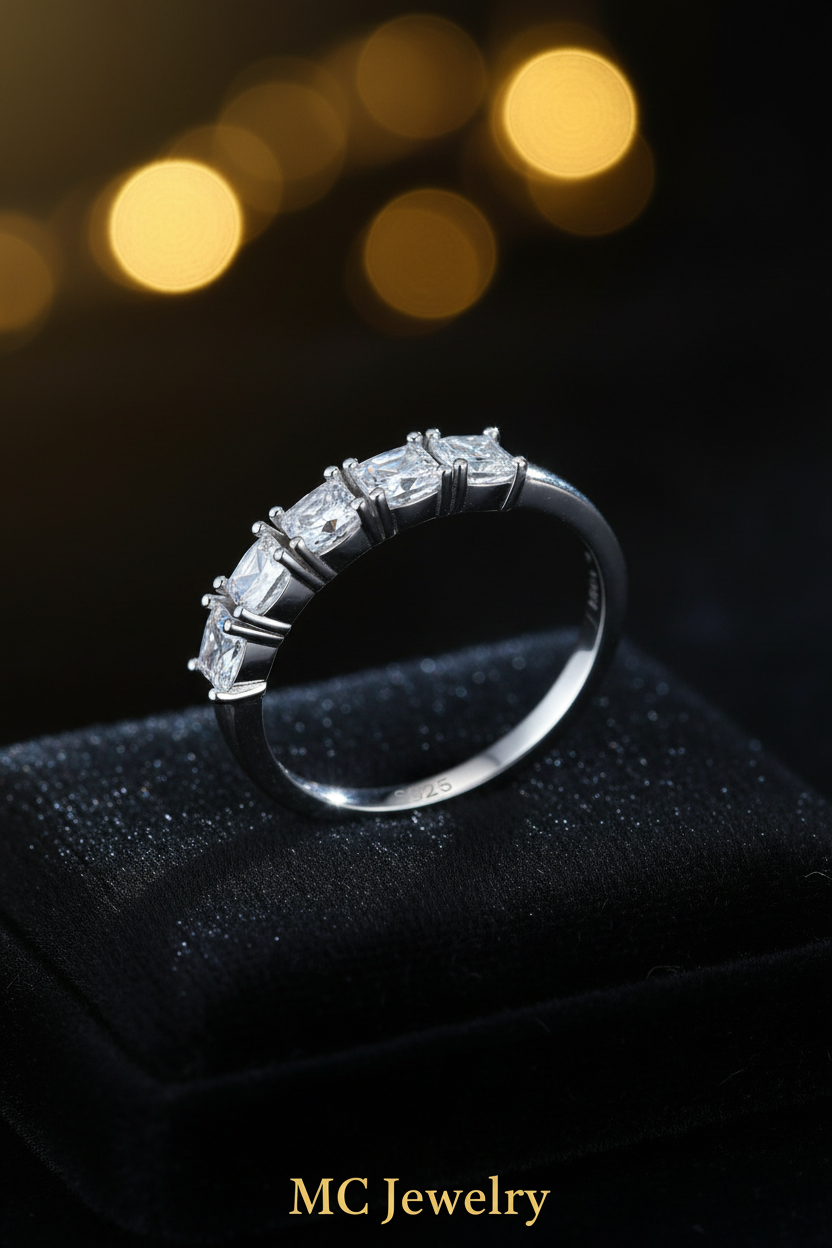 A silver-toned ring with a row of baguette-cut clear gemstones, displayed on a clear acrylic block against a dark blue reflective background.