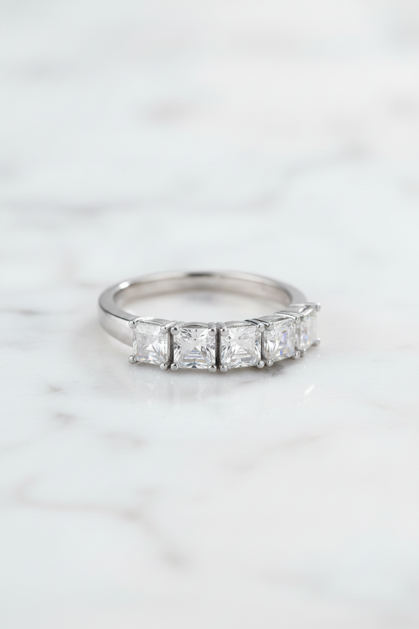 A silver-toned ring with a row of baguette-cut clear gemstones, displayed on a clear acrylic block against a dark blue reflective background.