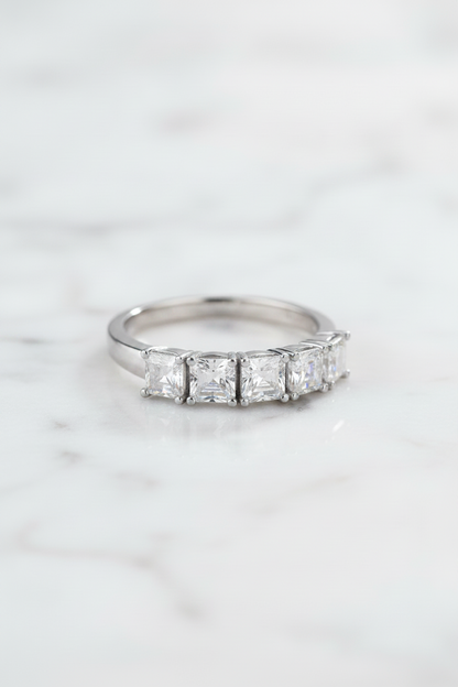 A silver-toned ring with a row of baguette-cut clear gemstones, displayed on a clear acrylic block against a dark blue reflective background.