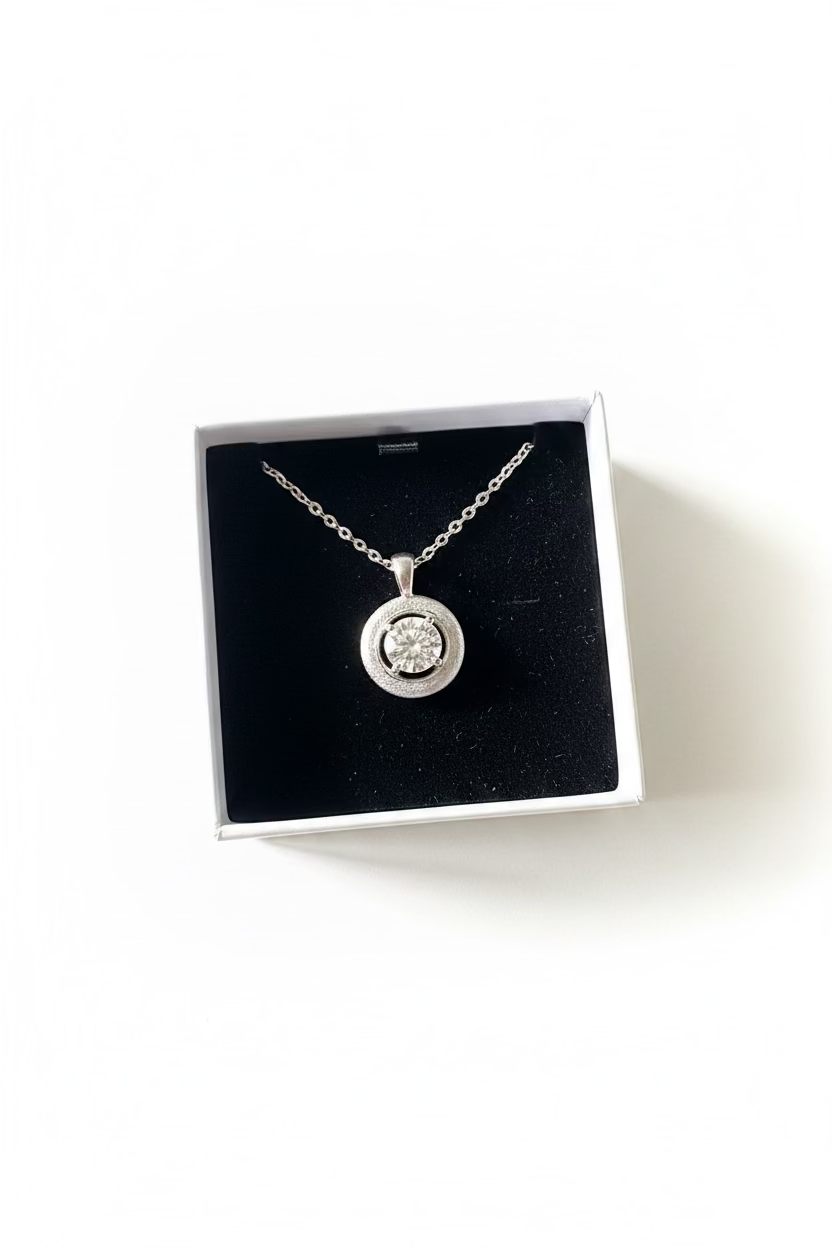 A silver-toned round pendant necklace with a clear central gemstone, displayed inside an open white jewelry box with a black 