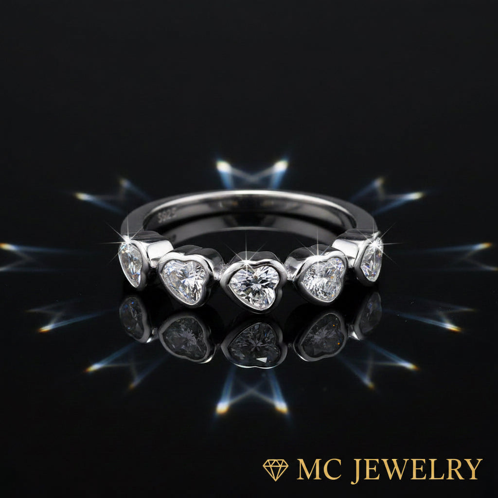 A silver band ring featuring five heart-shaped clear gemstones, displayed on a reflective black surface with starburst light effects. The MC JEWELRY logo is visible in the bottom right corner.