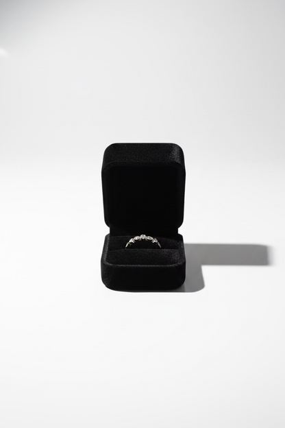 A thin gold band ring with a small central clear gemstone, displayed inside an open black velvet ring box 