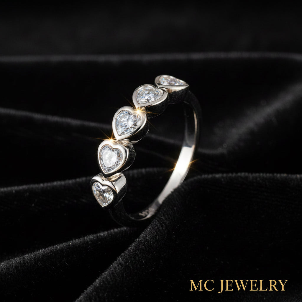 A silver band ring featuring five heart-shaped clear gemstones in a vertical row, displayed on a black velvet fabric background.