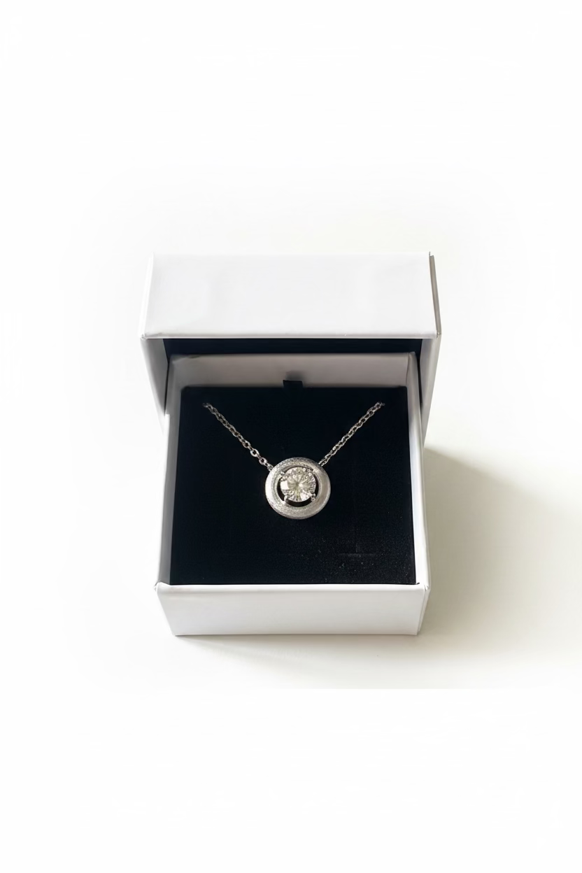 A silver-toned round pendant necklace with a clear central stone, displayed inside an open white jewelry box with a black velvet insert 